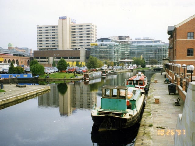 Canal Wharf at the Canal Basin looking in the direction of Victoria Bridge. Two barges are moored in front of Quayside House, right, opposite the Canal Basin, left. The boat in the foreground is The Horbury and acts as storage and supplies for the Kirkstall Flyboat moored behind it. The Kirkstall Flyboat is a purpose-built pleasure boat run by Yorkshire Hire Cruisers Ltd. It is equipped to take private parties of up to 50 people on cruises along the canal and River Aire. On the left a train can be seen crossing over the dark arches, about to enter the City Station. The large building in the background left is the Hilton Hotel in Neville Street.
