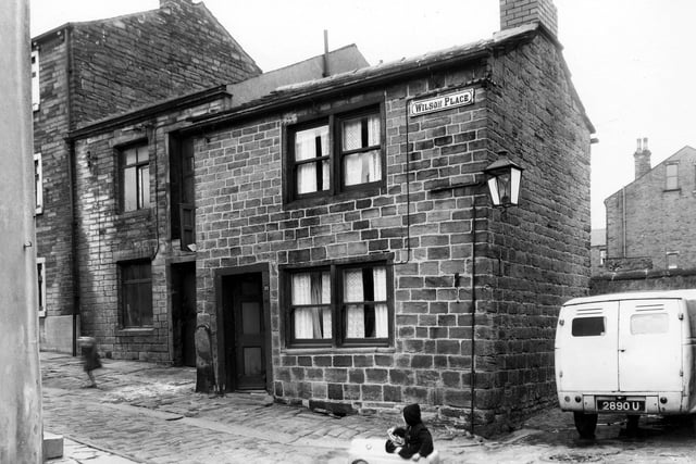 A view of Wilson Place, an enclave of old stone built terraced properties situated off Lower Town Street. The property on the left which is set back is the workshop of E. Dixon & Son, Funeral Directors and Wholesale and Retail Timber Merchants. Adjacent to it, right, is number 11 Wilson Place. A stone with a rounded top stands beside the door. A child in the foreground is negotiating a pedal car over the cobbles. Houses in Spring Terrace can be seen in the background, right. Pictured in March 1960.