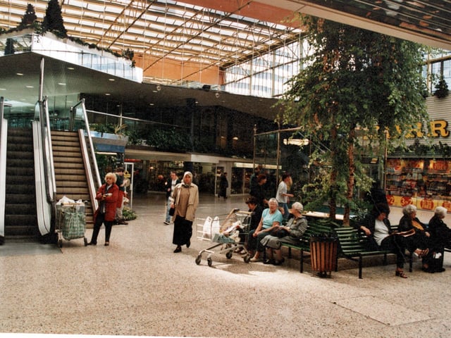 People sit on benches around artificial trees. Steps and an escalator are on the right. Morrisons is in the background.