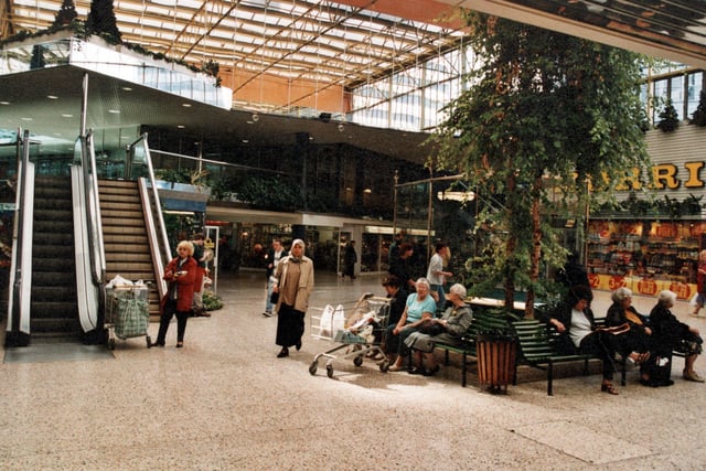 People sit on benches around artificial trees. Steps and an escalator are on the right. Morrisons is in the background.