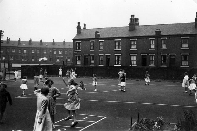 Another view of pupils of various ages at play in the playground of Burton House in Burton Avenue. The girls in the foreground are enjoying a game of hopscotch on a grid marked out on the surface of the playground. Behind the playground wall is the odd-numbered side of Dobson View (off Burton Avenue), a street of red brick back-to-back terrace homes. These are back-to-back with the even-numbered side of Dobson Terrace. The view also looks across Burton Avenue, and the grounds of Cockburn High School, to Back Fairford Place, background left. Burton House was originally a private residence, built in 1791.