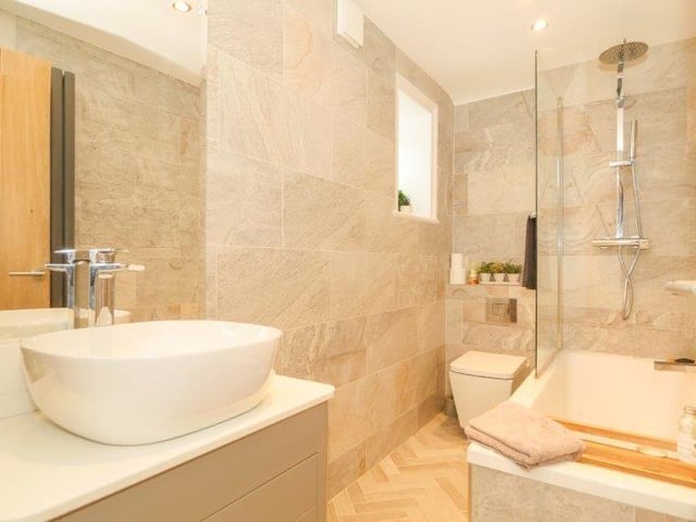 The bathroom features a luxurious three piece suite and includes an overhead mains powered rainfall shower, vanity style sink unit, wall mounted mirror and fully tiled walls and flooring.