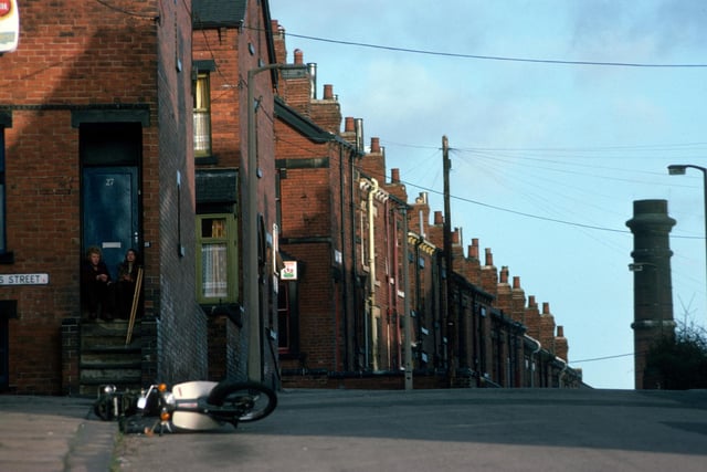 A view looking north along Woodhouse's Pennington Street in January 1976. The junction with Lucas Street is on the left, followed by Back Lucas Street and Lucas Place, after which the road continues down the hill as Back Burchett Grove. Two people sit on the steps in front of the door of no. 27 Lucas Street, while a motorbike lays on its side in the foreground. The top of the chimney of the Meanwood Road Refuse Destructor can be seen in the background on the right.