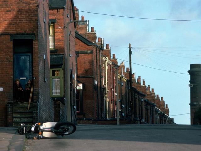 A view looking north along Woodhouse's Pennington Street in January 1976. The junction with Lucas Street is on the left, followed by Back Lucas Street and Lucas Place, after which the road continues down the hill as Back Burchett Grove. Two people sit on the steps in front of the door of no. 27 Lucas Street, while a motorbike lays on its side in the foreground. The top of the chimney of the Meanwood Road Refuse Destructor can be seen in the background on the right.