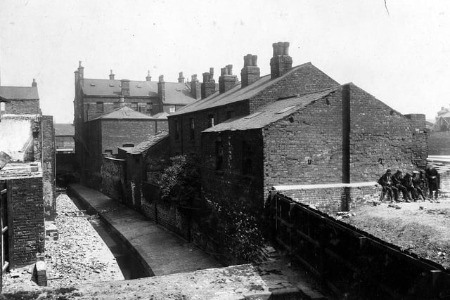 Lady Beck is on the left, the view is looking towards Lady Lane in June 1929. To the right are the rear views of houses which were St Peter's Court. St Peter's Street can just be seen on the right edge. The Marquis of Granby public house was situated at the corner of the two roads (not in view). A group of men are sitting on the wall. In the foreground is a slab bridge over the beck which connected Millgarth Street (to the left) to Wray's Yard (right, which is here blocked off by a wooden fence).