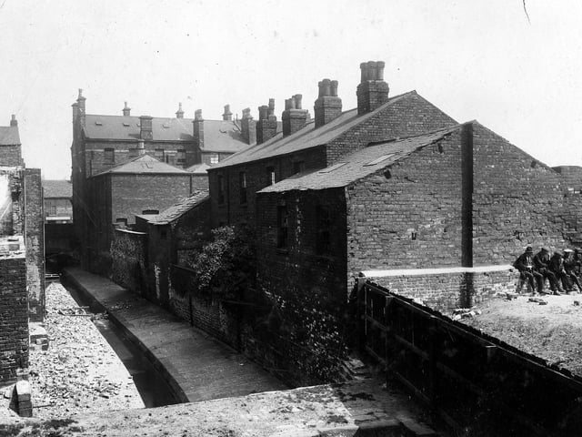 Lady Beck is on the left, the view is looking towards Lady Lane in June 1929. To the right are the rear views of houses which were St Peter's Court. St Peter's Street can just be seen on the right edge. The Marquis of Granby public house was located at the corner of the two roads (not in view). A group of men are sitting on the wall. In the foreground is a slab bridge over the beck which connected Millgarth Street (to the left) to Wray's Yard (right, which is here blocked off by a wooden fence).