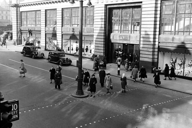 View of Lewis's (Leeds) Ltd. department store on The Headrow. Cars are parked outside. Pedestrians cross the road. Graffiti can be seen on Wade Lane. A double lampost, traffic lights and ladders are visible. Pictured in March 1949.
