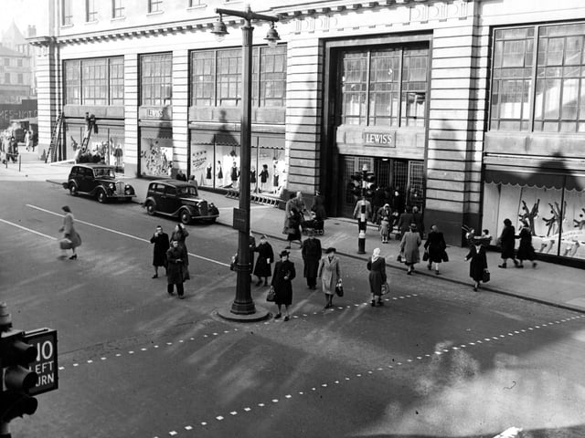 View of Lewis's (Leeds) Ltd. department store on The Headrow. Cars are parked outside. Pedestrians cross the road. Graffiti can be seen on Wade Lane. A double lampost, traffic lights and ladders are visible. Pictured in March 1949.