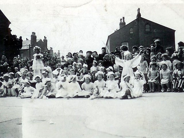 Children and spectators at an unknown event, possibly a May Day celebration, taking place in the school yard of Rowland Road Primary School. Many of the children are dressed in white with floral decorations, and behind them other children are seated on chairs, some in costume and wearing hats. At the rear is an audience of parents and carers.