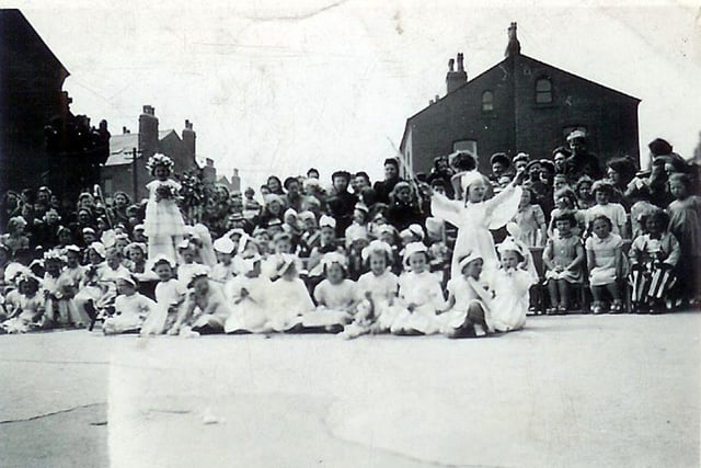 Children and spectators at an unknown event, possibly a May Day celebration, taking place in the school yard of Rowland Road Primary School. Many of the children are dressed in white with floral decorations, and behind them other children are seated on chairs, some in costume and wearing hats. At the rear is an audience of parents and carers.
