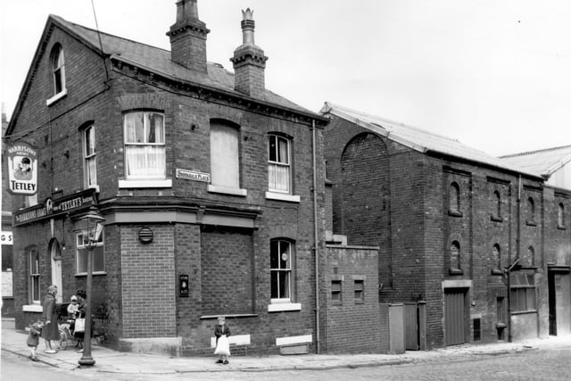 A view of the Harrison's Arms on the corner of Brookdale Place on Malvern Road. Two mothers stand chatting one with a coach built pram and a toddler perched on top. A small boy patiently waits round the corner clutching a large bag. Works premises can be seen on Brookdale Place.