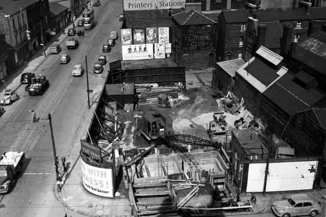 An aerial view of construction work on a sewer at the corner of Meadow Lane and Great Wilson Street in April 1955. A crane has its arm over the pit. Advertisements for Cadbury's Chocolate, Guinness, Chivers Jellies and others are visible. A painted sign for Wildblood & Ward, printers and stationers, can be seen.
