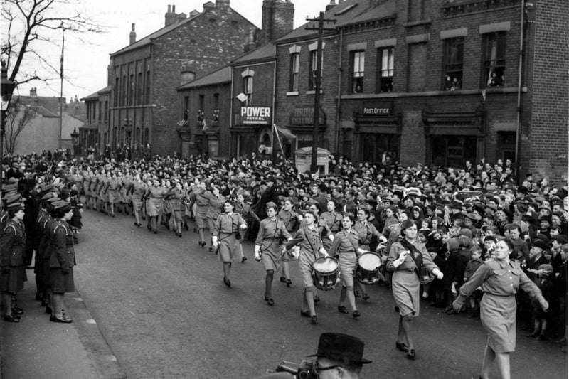 Parade on Rothwell's Marsh Street of The Royal Women's Junior Air Corps on the occasion of the visit of H.R.H. Princess Mary (The Princess Royal) for Salute the Soldier Week. Children and Adults line the street and onlookers look out from upstairs windows of businesses. The shops that are featured are The Post Office and Lesley Pyle, Cycle dealer.