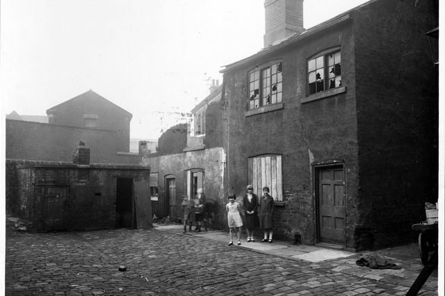 Chadwick Court off Bowman Lane in October 1935. There are two buildings visible : a derelict house which adjoins a collapsed house. On the left is a one-storey building possibly a lavatory, or storage area. To the far left of the photo, the wheels of a wooden cart can just be seen. There are several children watching the photographer.