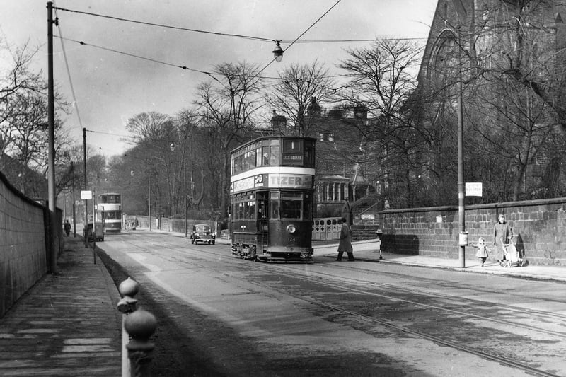 Headingley Lane by the junction with Cumberland Road in March 1956. Headingley Hill Congregational Church is seen on the right. Two trams are among the traffic on the road; no. 124 in front is on route no. 1 to City Square. A woman with a young child and a pushchair is walking past the bus stop on the right.