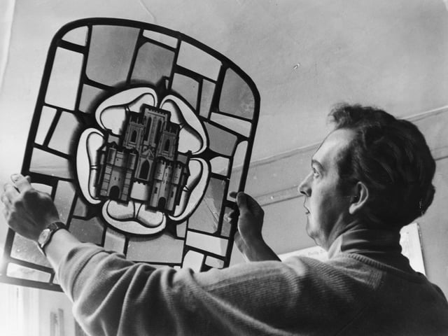 August 1970 and Sep Waugh holds up to the light a panel of a stained glass window he made for The Three Tuns Inn on Coppergate in York.