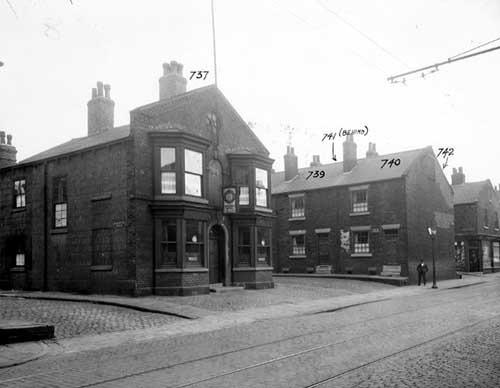 Balm Road in September 1939. The Railway Hotel is set back from road. Brick facade has double bay windows each side of door. Above the door is a sign for the hotel with the BYB symbol for Bentley's Yorkshire Beers. The left side of the hotel is cement rendered with street sign for Hardwick Street. To the right is Balm Street with man and gas lamp at end. Beyond can be seen a shop on the corner of Wright Street. In the foreground, the cobbles and tram lines of Balm Street.