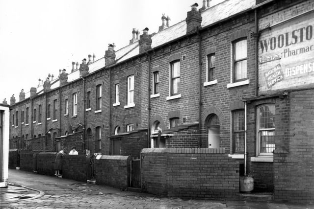 The rear of through terraced houses facing onto Devon Street. Every two houses there is a small lean-to building housing a separate toilets for each house. Washing lines extend across the yards. Two women talk across the step of no 5. On the far right an advert for Woolstons Pharmacies Ltd, National Health Service Dispensary, 89 Pontefract Lane is painted on the first floor of the building.