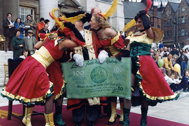 'Saloon girls' dressed in red, gold and green, present a 100 dollar bill to the Lord Mayor of Leeds, councillor Eric Atkinson MBE. On the occasion of the 7th annual Lord Mayor's Parade. The girls were part of the float entered by Lewis's department store in The Headrow depicting an American Western steam engine. When the Lord Mayor's Parade reached this point along the route in front of the Civic Hall steps the Lord Mayor received a salute from each participant. The floats were judged and Lewis's 'Fells Wargo Railway Co.' won the 'Lord Mayor's Award for the Best Overall Entry'. Collections and cheques presented went to Charities of the Lord Mayor's choice which were the Royal British Legion, Kidney Research Fund and National Association for mental health in 1980.
