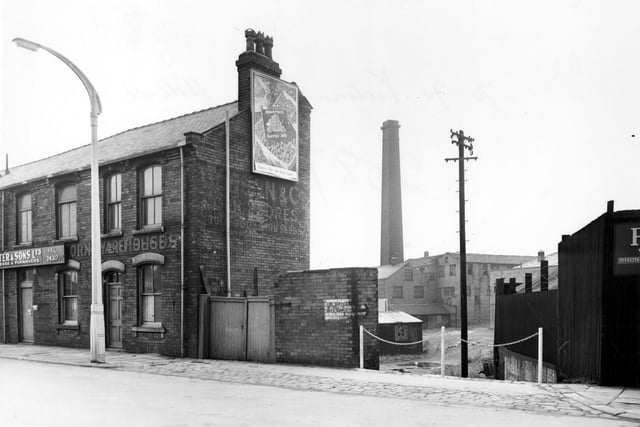 Victoria Road in August 1956. In background there is a derelict industrial building with tall brick chimney. Wooden fence with barbed wire to right of photo. Main building says in faded paint 'corn warehouse'. To gable end is a Rowntree Jelly advertising billboard. To the front is a new trade sign for an engineers and furnishers. A large new electric streetlight with concrete base stands in front of the row. A painted notice on a side wall indicates a fire service beck supply.
