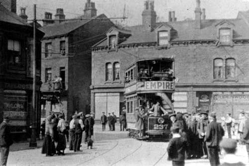 A tram at Morley Bottoms. This was the first tram from Morley to Leeds on a trial run. The route opened on 5th July 1911. The destination indicator says 'special car.' The tram is full of people while many have gathered to watch at the road side and from windows and balconies. Shops to the left include Taylor's and to the right Greenwoods.