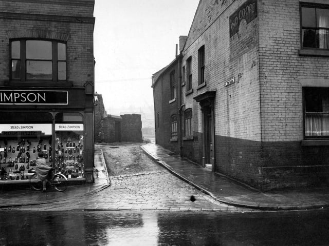 Looking south shows The Gang off Town Street in August 1952. Stead and Simpson shoe shop next door with a man on a bicycle looking in the window. A lamp is on the side of the building.