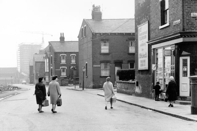 Green Lane looking south, showing the junctions with (from right) Hall Lane, Hawthorn Grove, Hawthorn Avenue, First Avenue and Second Avenue. A corner shop on the right (no. 21 Green Lane) displays an advertisement for Rhinds Radio and Television dealers of Tong Road on the wall. Three women are crossing the road, another is walking along the causeway on the right and a small boy is looking in the shop window. A tower block, probably Wortley Towers, is under construction in the background.