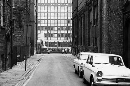A view down Alexander Street past the war memorial, over Victoria Gardens and over The Headrow to the Eagle Star Insurance Group building. On the left is the rear of buildings originally built in 1847 as offices and warehouses for the cloth industry, today this is the Henry Moore Centre for Sculpture. On the right is the side wall of the Municipal Buildings, now Leeds Art Gallery.
