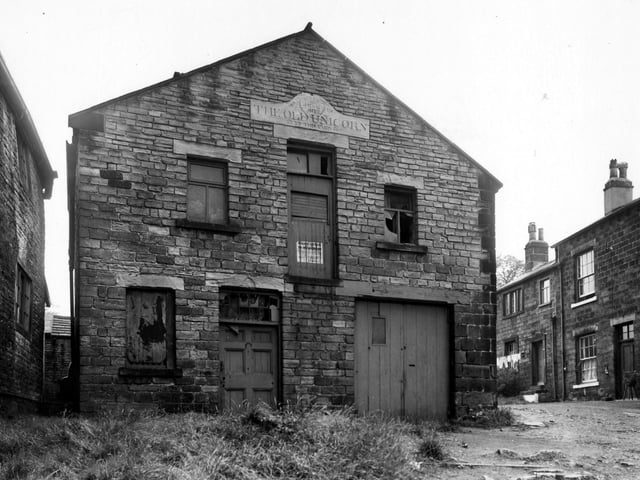 Stocks Hill in August 1956. Above, two windows to each side of a wooden loading bay, which has a police notice offering a ten shilling reward. Above this is an ornate carved stone for the Old Unicorn (up this yard), dated 1877. To the right is a row of stone cottages with washing outside one of them.