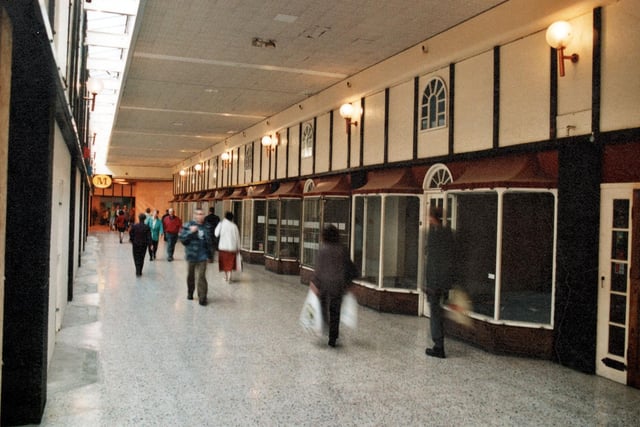 Morrisons is on the left with empty shop units on the right. The shops have arched, glass doorways and arched windows above. Lights hand off the wall and pedestrians are shown.