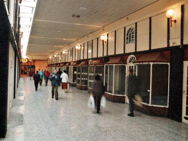 Morrisons is on the left with empty shop units on the right. The shops have arched, glass doorways and arched windows above. Lights hand off the wall and pedestrians are shown.