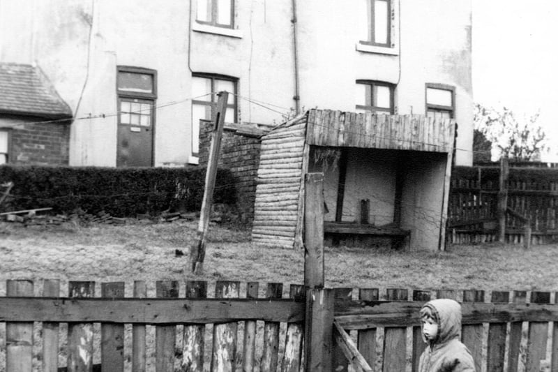 Hey Beck Lane in April 1967. A home-made wooden shelter with a bench seat stands on the grassed area. In the foreground there is a wooden fence topped with chicken wire. In front of the fence a small boy stands in a quilted, hooded anorak.