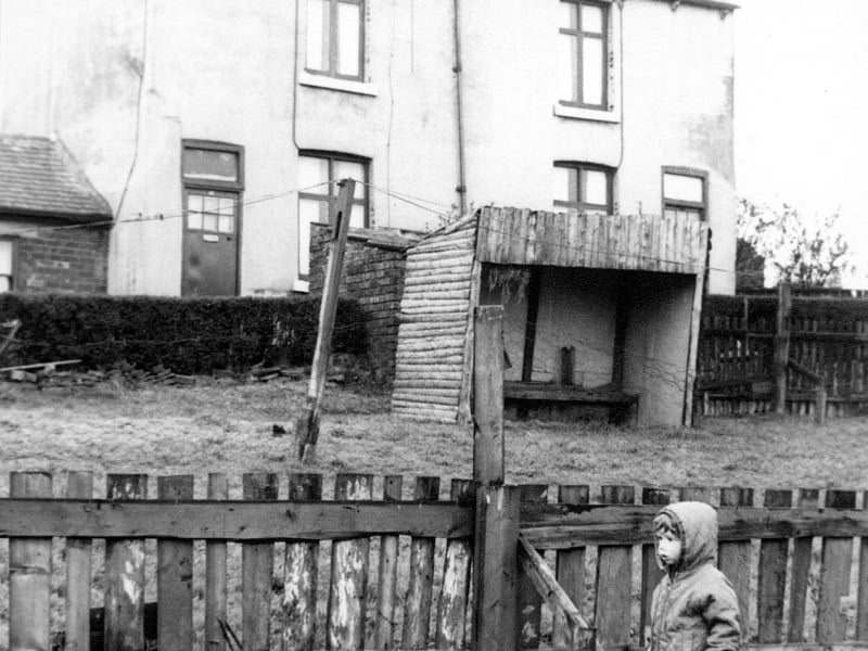 Hey Beck Lane in April 1967. A home-made wooden shelter with a bench seat stands on the grassed area. In the foreground there is a wooden fence topped with chicken wire. In front of the fence a small boy stands in a quilted, hooded anorak.
