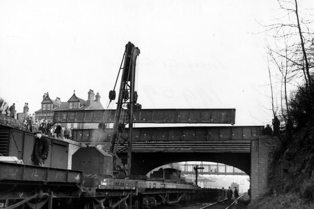 Construction work on Cross Gates Bridge on Station Road over the Leeds-York railway in January 1955. Onlookers and workmen watch as a crane lifts a girder into place. A footbridge, Cross Gates Station and the Station Hotel are in the background.