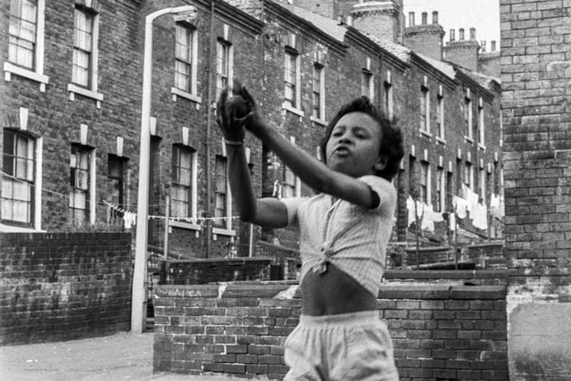 A young girl appears to be just catching a ball whilst playing out on Queen's Road. In the background is Hartwell Street, a back street which has on either side the back yards of the through terraces of Hartwell Place (left) and Hartwell Road (right).
