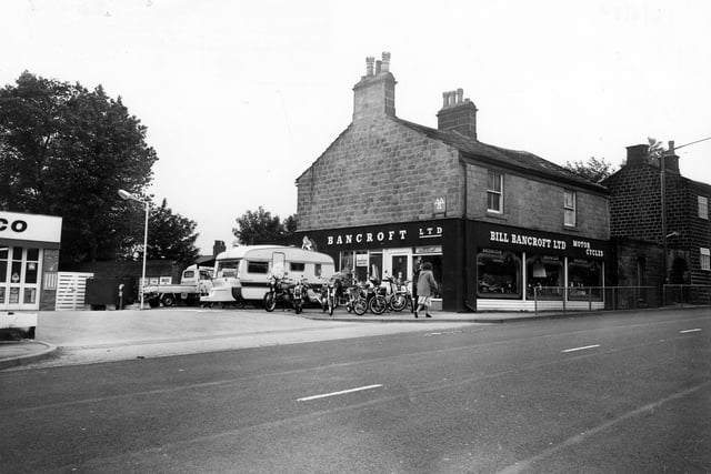 Bill Bancroft Ltd. Motor Cycles on New Road Side in Horsforth. The adjoining property is number 181 New Road Side. Various motor cycles are on diplay in the windows of the showroom and there are more parked outside alongside a caravan. The firm advertises as a Honda Specialist. To the left there is an Amoco Service Station.