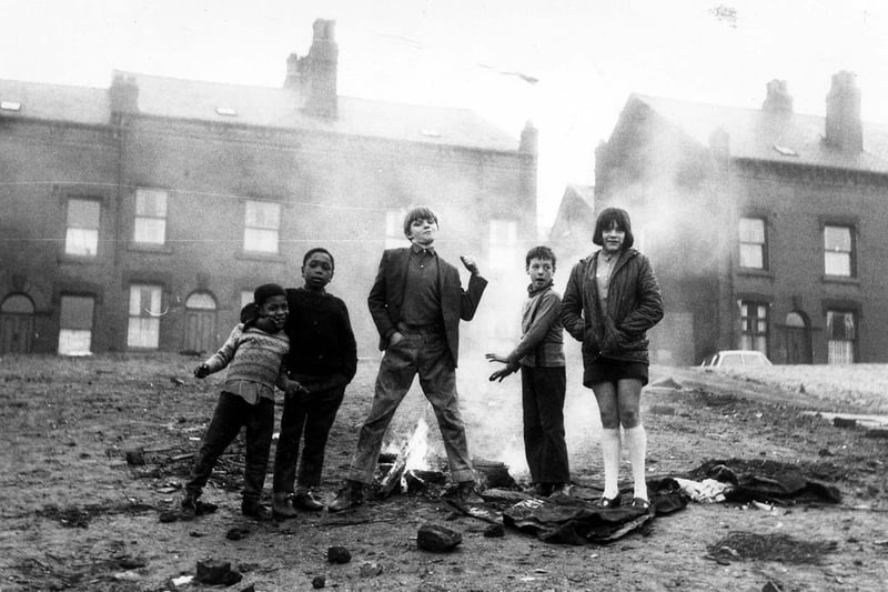 'Butch and Mates', shows a group of children of varying ages pictured as they play out on an area of cleared land in front of rows of terraced houses located in Servia Hill in 1970. They have lit a small "camp fire" which is blazing away behind them.