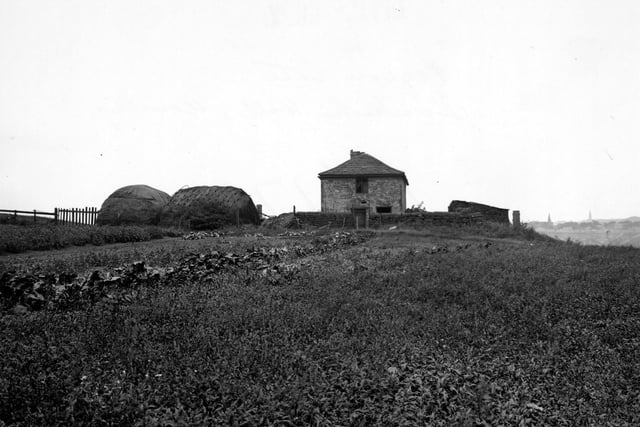 The old pigeon cote on Dunkirk Hill. The house has a stone built wall around it next to which are two large bales of hay. A field covers most of the foreground. On the right in the distance houses and church spires can be seen. Pictured in August 1949.