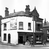 On the left edge of this view from  May 1965 is the single front of number 4 Strawberry Lane. Two shops on Tong Road follow to right, with a Dutch gable style roof above the top two windows. To the left, painted white, is number 170, a second hand furniture shop with a selection of cupboards outside. A blackboard with a chalked price list hangs next to the door. On the side wall is a billboard advertising salad cream with the slogan 'Heinz of course'. Number 168 follows to the right. This is Fred Brown's off-license and tobacconists.