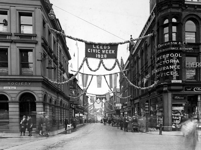 View looks from Albion Street onto a richly decorated Guildford Street. On the left is the Leeds and Holbeck Building Society with the Liverpool Victoria Insurance Offices and the Chocolate Bar beneath on the right. Banners and bunting are draped across the street along with the flags of many European countries.