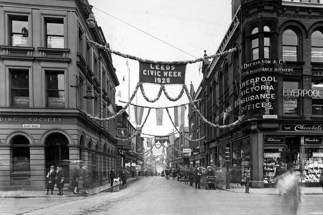 View looks from Albion Street onto a richly decorated Guildford Street. On the left is the Leeds and Holbeck Building Society with the Liverpool Victoria Insurance Offices and the Chocolate Bar beneath on the right. Banners and bunting are draped across the street along with the flags of many European countries.