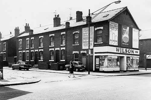 May 1967 and in view is Kirkstall Road with Cardigan Avenue on the left. A campaign poster for council election is on the wall, it reads 'Frank Marshall's team will get Leeds moving. Vote Conservative. Newton for Kirkstall'. On Kirkstall Road is a shop selling wallpaper, paint and other decorating supplies.