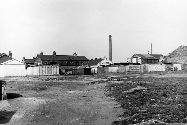 The waste ground on the corner of Compton Road and Florence Street in May 1953. There are several sheds and what looks to be a custom made van in the centre. To the right are two brick buildings, one with an advertising hoarding. The other building has a pile of timber leaning against it. In the distance are three rows of terraced brick houses and to the right of these is the Western cinema.