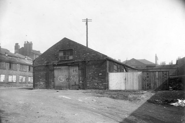 A derelict brick building with a large wooden gate, with graffitti on. The building is called Portland Gate. In the foreground lies some waste ground. To the left are some more derelict buildings. To the right is a wooden fence and wooden gate. Pictured in August 1929.