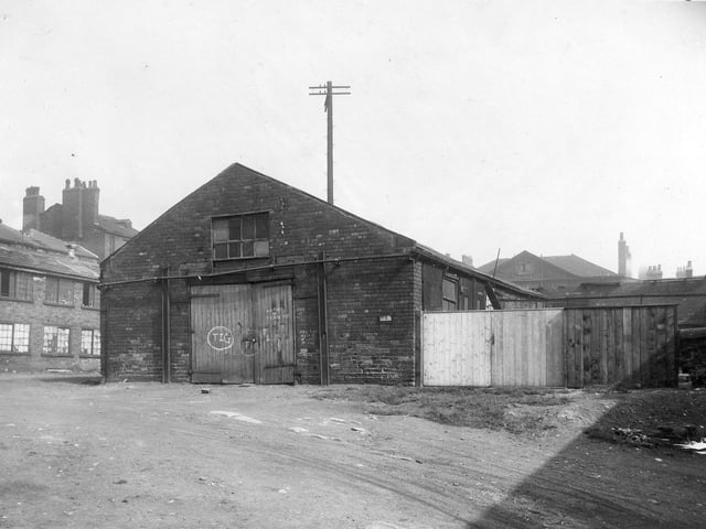 A derelict brick building with a large wooden gate, with graffitti on. The building is called Portland Gate. In the foreground lies some waste ground. To the left are some more derelict buildings. To the right is a wooden fence and wooden gate. Pictured in August 1929.
