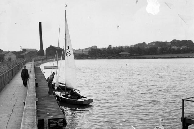 A view of Yeadon Tarn in 1955. On the left is a walkway and board walk for Yeadon Sailing club. Yachts and crew can be seen. The remains of waterside mill are visible, the large building which is next to the sail is called Ingots building and still stands, now headquarters of Headrow Housing. This is looking towards Cemetery Road.
