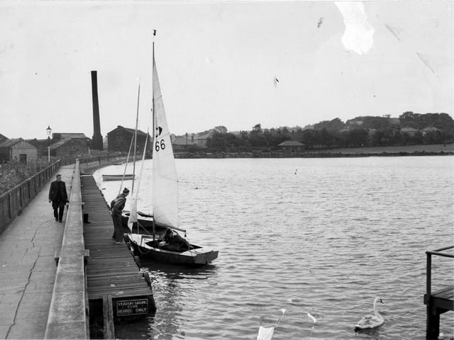 A view of Yeadon Tarn in 1955. On the left is a walkway and board walk for Yeadon Sailing club. Yachts and crew can be seen. The remains of waterside mill are visible, the large building which is next to the sail is called Ingots building and still stands, now headquarters of Headrow Housing. This is looking towards Cemetery Road.
