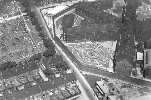An aerial view of of Armley Gaol in 1937. It was built as Leeds Borough Gaol and completed in July 1847 at a cost of £43,000. The buildings were designed by architects Perkin and Backhouse. The road from top to bottom across the centre is Hall Lane and New Wortley Cemetery is at the left edge from the middle to the top. The prison is surrounded by a high wall and wihin the boundaries there is an exercise yard. Inmates can be seen walking the circular paths. The rows of terraced houses, bottom left, are Hawthorn Place and Hawthorn Terrace. Behind the prison Winchester Road runs from the junction with Hall Lane.