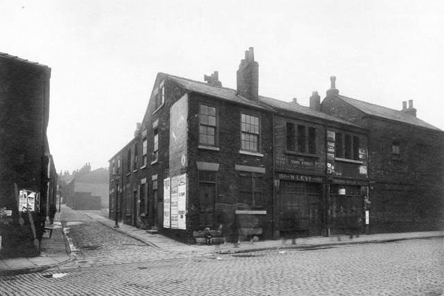 Looking from York Street along the length of Plane Street in the city centre in June 1932.. This shows the empty shops and houses just prior to demolition. Both streets are cobbled with dressed stone blocks. Two gas streetlamps are visible on the pavement. A small child is sitting on the steps of the corner building.