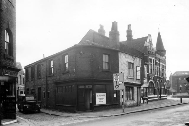 On the left edge of this view, the doorway of Lake's corner, a grocers run by Frederick Lake can be seen on Holbeck Lane. The entrance to Stead Street follows to the right where a van for John Cooke and Son of Huddersfield is parked. This firm were concrete engineers and had works on Coleman Street nearby. Continuing right is C. Tobin's licensed betting office at number 77. Outside this shop is a road sign directing motorists along Wortley Lane to Huddersfield and Halifax and along Spence Lane to Armley.  Pictured in March 1965.